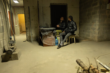 View of a basement with caucasian guy and black guy are sitting and having a talk near laptop...