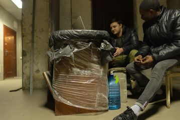 Bomb-shelter with caucasian guy and black guy sitting and having a talk near laptop during a war