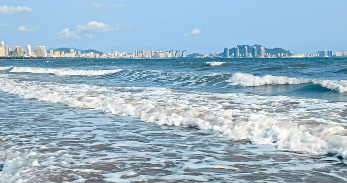Rolling waves and city skyline in Sanya, Hainan Island, China. Beautiful seascape. Yellow sand beach, azure sea and blue sky.