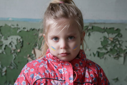 Portrait of little girl in ruined building with Ukraine flag painted on her face. Refugees, war crisis, humanitarian disaster concept.
