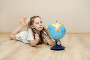 little cute girl lies on the floor, holds a magnifying glass in one hand, and holds the globe in three, carefully studying it