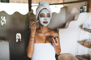 Asian female in white towel doing cosmetic procedures for face and thinking during nourishing moisturize with makeup brush, young attractive woman apply natural organic cream or alginate mask
