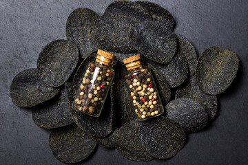 Black potato chips on a black background. A mixture of peppercorns in glass flasks next to chips. potato product