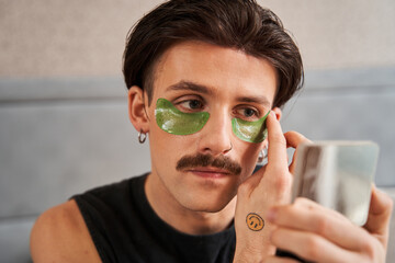 Brunette man with mustache sitting at the bed and applying eye patches