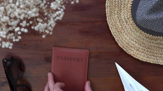 Flat lay travel concept video. Focus on passport in centre of frame. Persons hand takes passport and checks in before replacing it in shot. Holiday items surround frame edges.