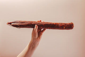 Kulen in hand on beige background. Meat sausage made of minced pork with red paprika, traditionally produced in Croatia and Serbia.