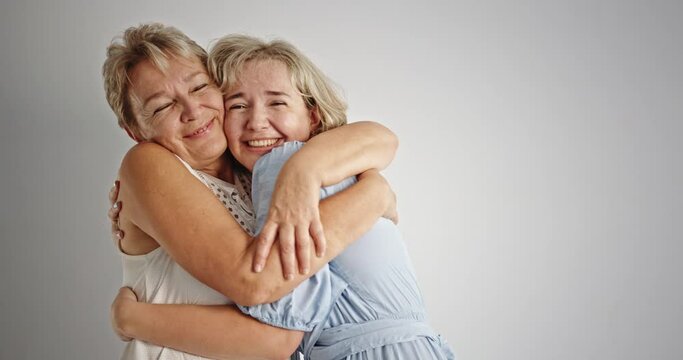 Mother and daughter hugging each other. Cheerful mature mom smiling and looking at camera while embracing adult daughter against gray background
