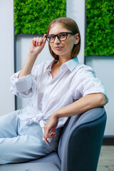 Obraz premium A young woman in glasses with a smile on her face, she is in a white shirt and jeans sitting on the sofa