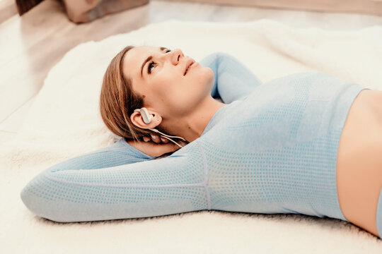 Top view portrait of relaxed woman listening to music with headphones lying on carpet at home. She is dressed in a blue tracksuit.