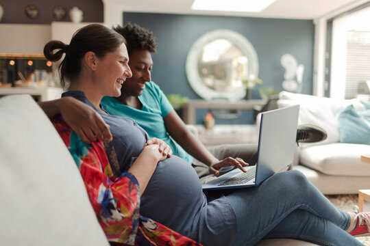 Pregnant couple using laptop on living room sofa - Powered by Adobe