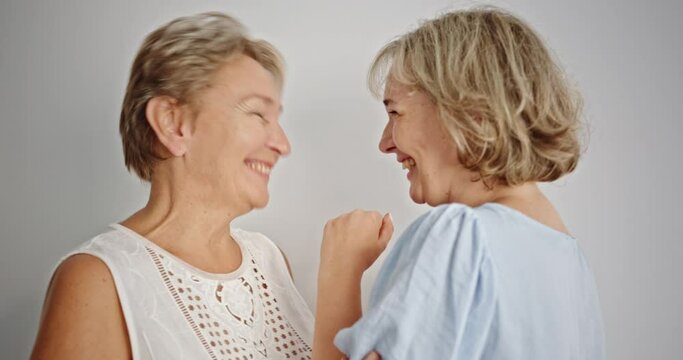 Mother and daughter gossiping and laughing. Blond adult daughter whispering secret to ear of mother then laughing cheerfully against gray background