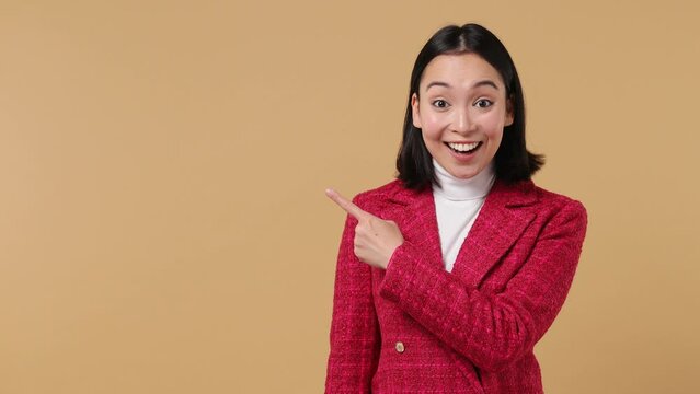 Promoter young woman of Asian ethnicity 20s wears red jacket pointing fingers hands aside on workspace copy space mockup promo commercial area isolated on plain pastel beige background studio portrait