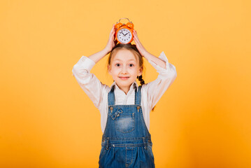 Isolated on yellow, attractive caucasian child in blue jeans with long bright hair hold alarm clock