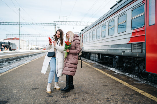 two girls on the station platform with flowers happily looking into a smartphone