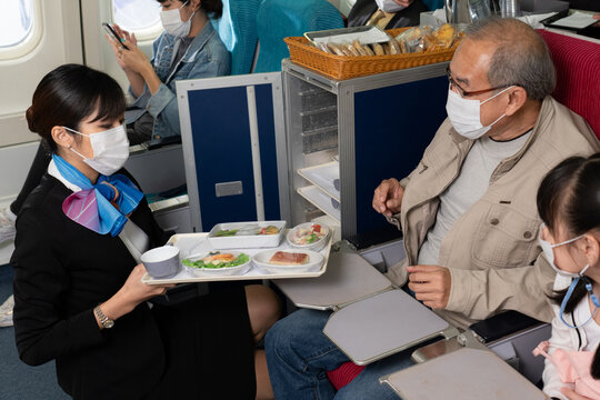 Asian Female Flight Attendant Serving An Airline Meal To Asian Senior Male Passenger And His Granddaughter, Placed On A Seat Tray Table. Everyone In The Plane Wearing A Face Mask To Protect Covid-19.