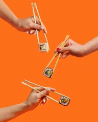 Poster Women's hands hold sushi rolls with sticks. Orange background. Creative concept © Max
