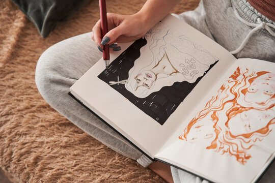 Relaxed female artist sitting at the sofa and drawing sketches at her notebook at home