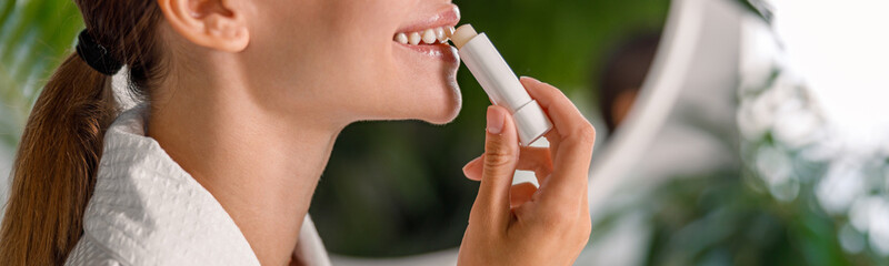 Side view of smiling woman in bathrobe applying lip balm, taking care of her lips, standing in...