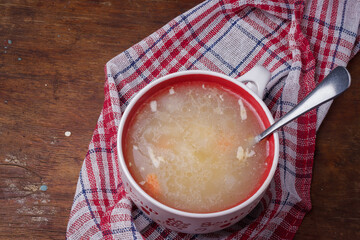 Poultry soup, homemade fat chicken broth with vegetables in red bowl on kitchen towel, wooden rustic table, close up. Healthy food for immune system