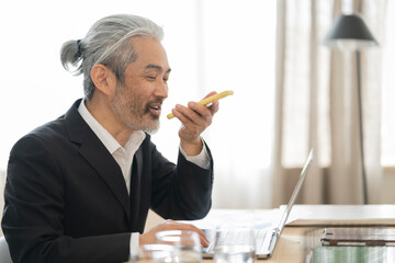 gray haired mature asian man with ponytail office worker sitting with computer talking on mobile...