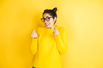 Young beautiful woman wearing casual sweater over isolated yellow background doing money gesture with hands, asking for salary payment, millionaire business - obrazy, fototapety, plakaty