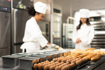 Selective focus of many empty baking bread loaf pans placed near two trays of 30 chicken eggs and a tray of baked breads on a table where blurred two female bakers are kneading dough in a kitchen.