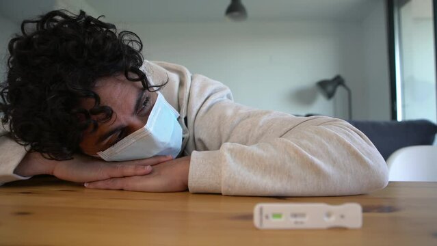A sad caucasian man makes a rapid home test device for Covid19 virus with a positive result and holding his head on hand at wood table. Antigen cassette of pcr to new Omicron variant virus.