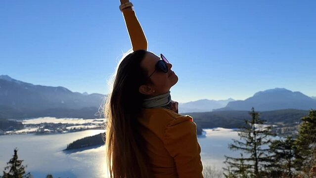 A woman flipping her hair with a scenic view on Faak lake in Carinthia, Austrian Alps. Slow motion. The picturesque lake is surrounded by high mountains. City of Villach tourism. Happiness concept.