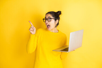 Young brunette woman working using computer laptop over yellow background amazed and pointing with hand and finger to the side
