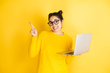 Young brunette woman working using computer laptop over yellow background smiling and pointing with hand and finger to the side
