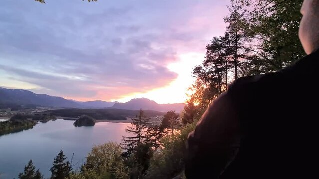 A woman watching the sunset with a scenic view on Faak lake in Carinthia, Austrian Alps. Sun down. The picturesque lake is surrounded by high mountains. City of Villach tourism. Happiness concept.
