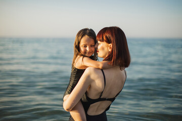 Family look concept. Happy mother in in black swimsuit with little daughter hugs her shoulders on beach. Beautiful red hair woman and her babe celebrating mother's day by the sea.