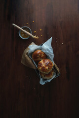 A flat lay image of hot cross buns set against a dark background. Easter baked goods in a baking tray with a marmalade jam glaze and pastry brush in shot. Copy space available