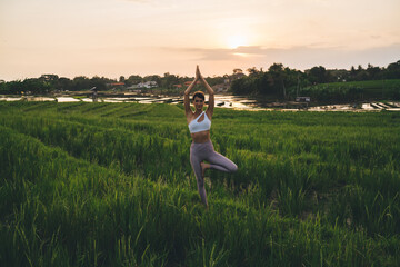 Full length portrait of Caucasian fit girl in sportive tracksuit standing in tree pose and looking at camera during summer time for visiting rice fields of Indonesia, woman posing and stretching