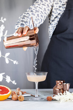 Real woman preparing Pink Lychee Cocktail in Champagne coupe glass surrounded by ingredients and bar tools