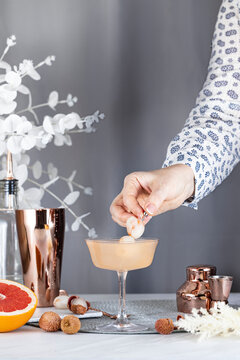 Real woman preparing Pink Lychee Cocktail in Champagne coupe glass surrounded by ingredients and bar tools