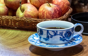 Small porcelain coffee cup, with blue floral pattern, and apples in a basket on a wooden table.