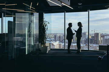 Fototapeta na wymiar Silhouettes of two employees team of businessmen, Asian man and woman are consulting and communicating, working in a modern office by the window