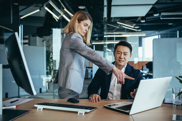Fototapeta premium happy coworkers discuss project strategy by looking and pointing at laptop pc computer monitor screen. multiethnic business team in the office. confident mature asian man explaining young female