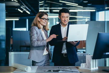 A team of multiracial business architects and designers discuss creating a project draft drawing by looking at laptop monitor screen a computer desk. Asian male and female developers together teamwork