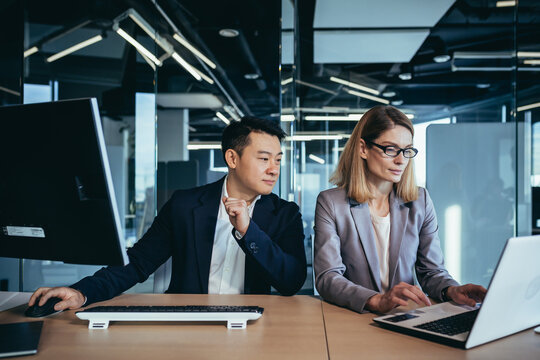 Happy Coworkers Discuss Project Strategy By Looking And Pointing At Laptop Pc Computer Monitor Screen. Multiethnic Business Team In The Office. Confident Mature Asian Man Explaining Young Female