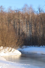 Snow - covered banks of a small forest river covered with ice .