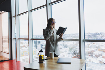 Experienced business woman holding a phone while standing in a modern office interior near a large window overlooking the cityscape. Female executive looks pleased after a successful meeting