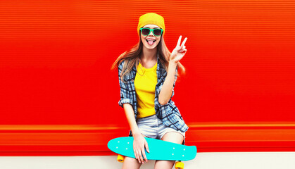 Portrait of stylish young woman model with skateboard wearing colorful yellow hat on red background