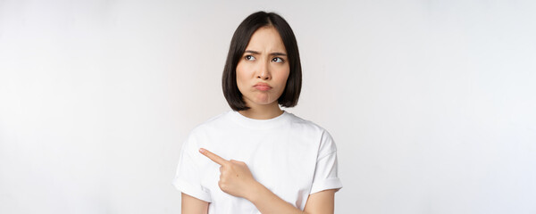 Angry and upset young asian woman pointing left, looking with regret or jealousy, feel unfair,...