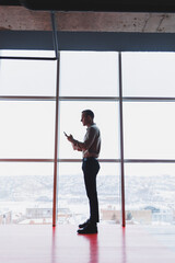 Fototapeta na wymiar A young executive is holding a phone while standing in an office interior and looking out a large window overlooking the city. Male manager with a phone is thinking about a new business project