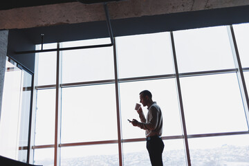 A young executive is holding a phone while standing in an office interior and looking out a large window overlooking the city. Male manager with a phone is thinking about a new business project