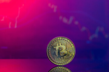 Close-up of a golden Bitcoin on a pink and blue reflective surface and the histogram of decreasing crypto in the background