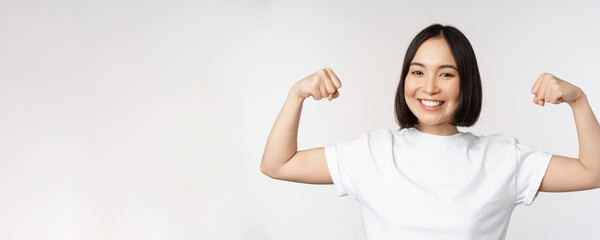 Fototapeta na wymiar Smiling asian woman showing flexing biceps, muscles strong arms gesture, standing in white tshirt over white background