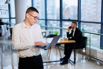 A young businessman man in glasses and a white shirt with a laptop in his hands stands in the office and works